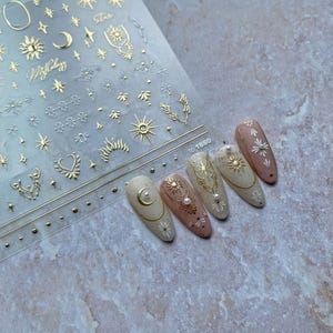 May include: A set of almond-shaped nail art designs. The nails are painted in neutral tones and adorned with gold celestial stickers, including suns, moons, and stars. Some nails feature pearl accents. A sheet of gold celestial stickers is visible in the background.