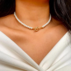 May include: A necklace featuring a single strand of uniform white pearls. The necklace is secured at the front with a gold-toned metal toggle clasp, which consists of a circular ring and a T-shaped bar, creating a central focal point.