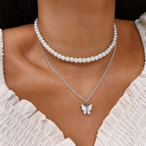 May include: A two-strand necklace. The top strand is made of white faux pearls. The lower strand is a silver chain with a small, white butterfly pendant. The necklace is worn over a white top.