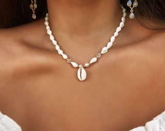 Puede incluir: Un collar de conchas blancas con pequeñas cuentas blancas y detalles de cristal transparente. El collar tiene una cadena dorada y un colgante de concha.