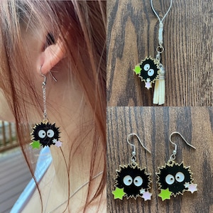 May include: Black and white cartoon character earrings with green and pink star accents. The earrings have a silver chain and hook. A matching charm with a tassel is also shown.