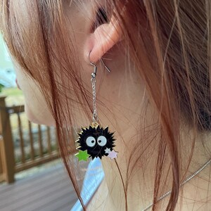 May include: A dangling earring featuring a black, round character with white eyes and a spiky outline. The earring has a silver chain and hook, with green and pink star accents.