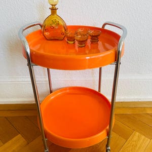 Puede incluir: Un carrito de servicio naranja de dos niveles con estructura cromada. La bandeja superior contiene una garrafa decorativa de vidrio ámbar con diseño floral y tres vasos pequeños. La bandeja inferior está vacía. El carrito tiene ruedas.