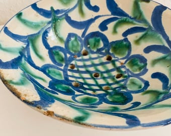 May include: A ceramic bowl with a white base and intricate blue and green floral patterns. The center features a grid design with several small holes. The rim has a slightly worn, rustic appearance.