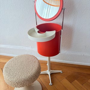 Peut inclure: Une coiffeuse orange vif dotée d'un miroir circulaire, d'un compartiment de rangement cylindrique et d'une étagère blanche en demi-cercle. L'unité repose sur une base blanche à quatre pieds. Un tabouret assorti au siège beige texturé est inclus.