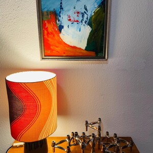 May include: An abstract painting in a gold frame hangs above a table with a lamp and candle holders. The lamp has a black base and a shade with orange, red, and brown stripes. Silver candle holders are arranged on the table.