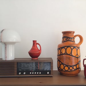 May include: A vintage wooden radio with a white ceramic lamp with a red and black stripe design, a red ceramic vase, a large orange ceramic vase with black circular designs, and a small red ceramic vase on a wooden surface.