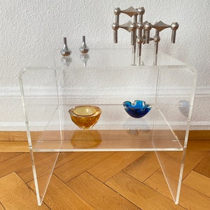 Puede incluir: Una estantería de acrílico transparente con dos estantes. El estante superior tiene tres candelabros de plata y un candelabro de plata. El estante inferior tiene un cuenco de vidrio azul y un cuenco de vidrio amarillo.
