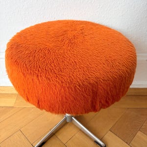 Könnte beinhalten: Ein runder Hocker mit einem leuchtend orangefarbenen Sitzpolster aus zotteligem Kunstfell. Der Sitz wird von einem silberfarbenen Metallgestell mit drei Beinen getragen, die von einem zentralen Punkt nach außen verlaufen und für Stabilität sorgen.