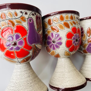May include: Three ceramic goblets with a speckled beige base and a dark brown rim. The goblets feature colourful floral and bird designs in red, purple, orange, and brown. The stemware has a unique, artistic appearance.