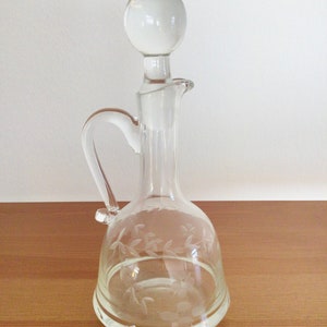 May include: A clear glass decanter with a round stopper and a floral etched design. The decanter has a handle and a wide base.