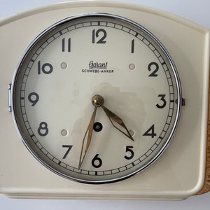 Puede incluir: Un reloj blanco con una esfera cromada y números negros. El reloj tiene una aguja horaria y una aguja minutera doradas. La esfera del reloj dice "Garant SCHWEBE-ANKER".