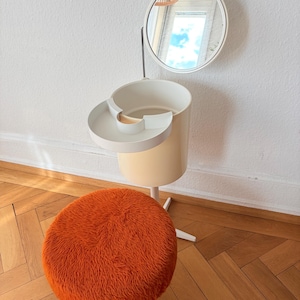 Könnte beinhalten: Ein Frisiertisch-Set bestehend aus einem runden orangefarbenen Hocker mit zotteligem Kunstfellbezug auf einem vierbeinigen Metallgestell und einer weißen zylindrischen Aufbewahrungseinheit mit Schwenktablett, Stange und rundem Spiegel.