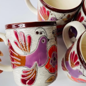 Könnte beinhalten: Set aus vier Keramikbechern mit weißem Hintergrund und bunten Vogel- und Blumenmotiven. Die Becher haben braune Ränder und Griffe.