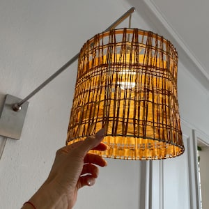 May include: A cylindrical wall-mounted lamp featuring a woven amber-colored fiber shade with a grid-like pattern. The shade is supported by a silver-toned metal arm that extends from a rectangular wall plate, with a light bulb visible inside.