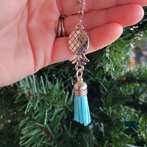 May include: A silver pineapple keychain with a blue tassel. The keychain is attached to a silver ring.