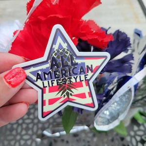 May include: A star-shaped sticker with a pineapple design and the text "All American Lifestyle". The sticker is on a background of red, white, and blue carnations.