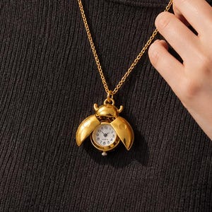 May include: Gold-coloured necklace with a beetle-shaped pendant. The pendant opens to reveal a small, round watch with a white face and black numerals. The necklace is worn against a black top.