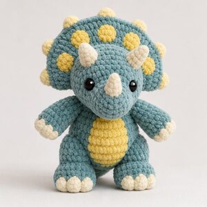 May include: A crochet triceratops plush toy featuring a textured blue body with a pale yellow belly, horns, and frill accents. The dinosaur has two large black safety eyes and a rounded head with a prominent frill decorated with yellow spots.