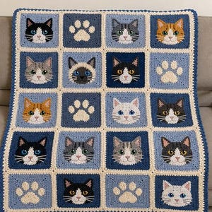 May include: A crochet blanket featuring a grid of square panels in shades of blue and cream. The panels display various cat faces with different coat colors and patterns, interspersed with cream-colored paw print motifs throughout the design.