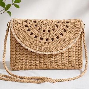 May include: A beige woven straw crossbody bag featuring a rectangular body and a semi-circular flap with a decorative openwork pattern. The bag includes a braided straw shoulder strap attached to the sides with small gold-tone metal rings.