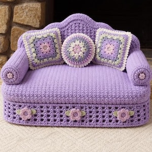 May include: A lavender crochet sofa featuring a textured seat and backrest, two square pillows with floral motifs, and one round central pillow. The base is decorated with three crocheted flowers, and the armrests are finished with small floral accents.