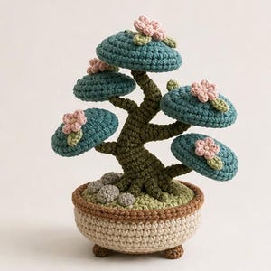 Peut inclure: Un bonsaï crocheté avec un tronc et des branches en vert olive texturé, surmonté de coussinets de feuillage bleu sarcelle arrondis ornés de petites fleurs rose pâle. L'arbre repose dans un pot crocheté beige rond avec une bordure marron.