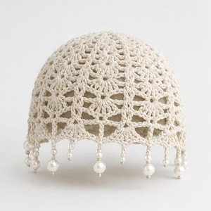 May include: A cream-colored crochet cap featuring an intricate, repeating floral-inspired pattern throughout the dome. The bottom edge is finished with a delicate fringe of dangling white faux pearls and small silver-toned beads.