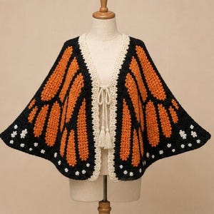 May include: A crochet capelet featuring a monarch butterfly wing pattern in vibrant orange and black yarn. The garment has a cream-colored border, a front tie closure with two tassels, and small white accents along the bottom edges.