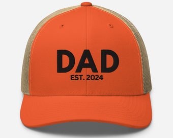 Op de afbeelding: Oranje en bruine truckercap met zwarte geborduurde tekst "DAD EST. 2024".