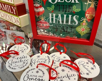 Op de afbeelding: Een verzameling witte, ronde kerstversieringen met rode linten. Elke ornament heeft zwarte tekst, waaronder zinnen als "Rabbi Teacher" en "Alpha and Omega". Een ingelijst bord met "Deck the Halls Y'all" op de achtergrond.