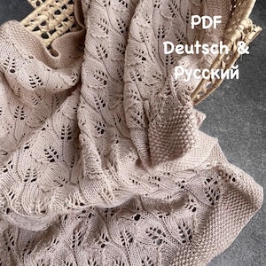 Könnte beinhalten: Eine hellbeige gestrickte Decke mit einem komplizierten Spitzen-Blättermuster und einem kraus rechts gestrickten Rand. Der Text PDF Deutsch & Русский ist auf dem Stoff sichtbar. Die Decke liegt über einem geflochtenen Korb.