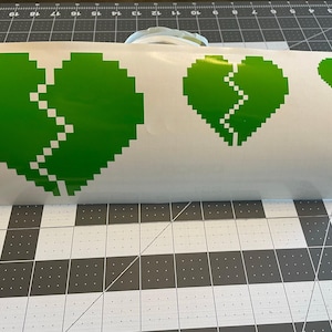 May include: A white sheet of vinyl with three green pixelated heart decals. The largest heart is broken in half with a jagged line. The two smaller hearts are also broken in half with jagged lines.