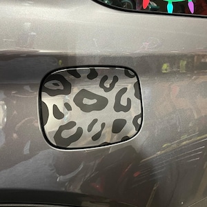 May include: A gray car fuel door cover with a black and gray leopard print design. The cover is rectangular with rounded corners and is set against a gray car body.