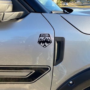 May include: A silver car with a black "Base Camp" sticker on the side. The sticker features a mountain range and a tent.