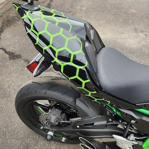 May include: Black motorcycle with a seat and rear tire visible. The motorcycle's rear section features a black and green honeycomb pattern. The exhaust pipes are silver. The motorcycle is parked on a concrete surface.
