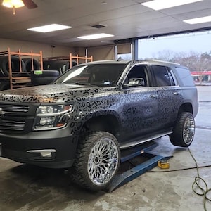 May include: A black SUV with a leopard print design on the hood and sides, parked on a blue lift. The vehicle has large chrome rims and tires. The background shows a tire shop with stacks of tires.