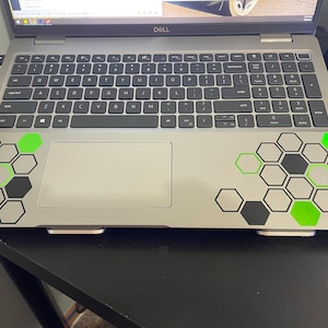 May include: A silver laptop computer with a black and green honeycomb design on the bottom half of the lid. The laptop is closed and has a Dell logo on the top of the lid.