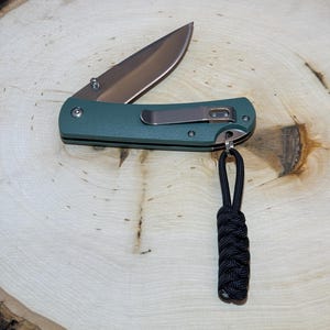 May include: A green pocket knife with a black paracord lanyard. The knife is closed and has a silver clip.