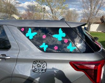 May include: A gray SUV with a rear window decorated with turquoise butterfly and pink flower decals. The decals are arranged in a symmetrical pattern on the window.