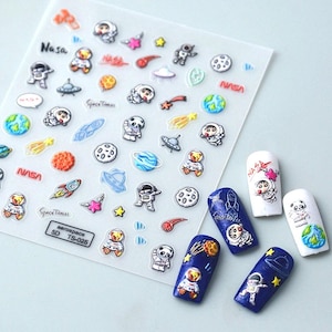 May include: A sheet of clear nail stickers with a space theme. The stickers feature astronauts, planets, stars, rockets, and the words "Space Travel" and "Aerospace".
