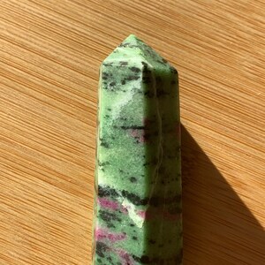 Puede incluir: Una punta de piedra preciosa verde, rosa y negra, también conocida como cristal, con forma facetada. La punta está sobre una superficie de madera.