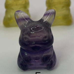 May include: A purple fluorite rabbit figurine with a glossy finish. The figurine is sitting on a white surface with the letter "E" printed below it.