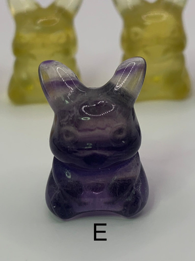 May include: A purple fluorite rabbit figurine with a glossy finish. The figurine is sitting on a white surface with the letter "E" printed below it.