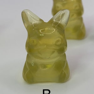 May include: Two translucent yellow gummy candies shaped like a cartoon character with large ears and a wide smile. The candies are sitting on a white surface. The letter "B" is printed below the candies.