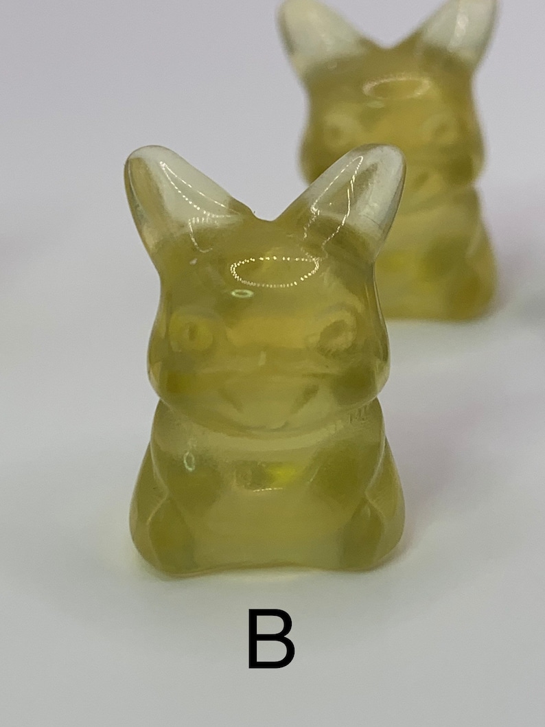 May include: Two translucent yellow gummy candies shaped like a cartoon character with large ears and a wide smile. The candies are sitting on a white surface. The letter "B" is printed below the candies.