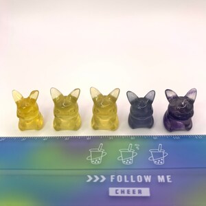 May include: Five small figurines of a cartoon character, possibly Pikachu, in different colors. The figurines are lined up in a row from left to right: yellow, yellow, yellow, purple, and purple. The figurines are sitting on a white surface in front of a ruler with a rainbow gradient.
