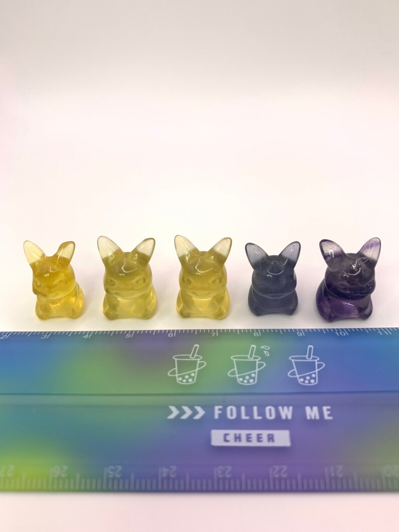 May include: Five small figurines of a cartoon character, possibly Pikachu, in different colors. The figurines are lined up in a row from left to right: yellow, yellow, yellow, purple, and purple. The figurines are sitting on a white surface in front of a ruler with a rainbow gradient.