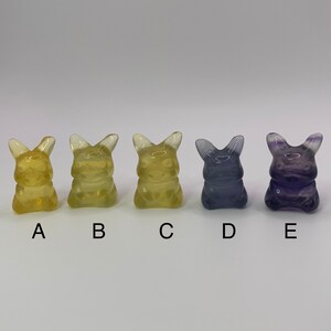 May include: Five small, clear, glass figurines in the shape of a cartoon character. The figurines are arranged in a row from left to right. The first three figurines are yellow, the fourth figurine is light blue, and the fifth figurine is purple.