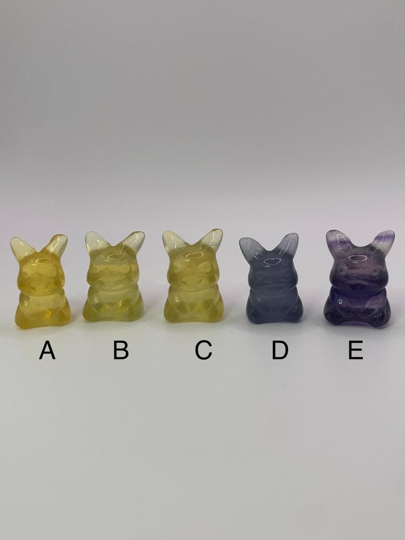 May include: Five small, clear, glass figurines in the shape of a cartoon character. The figurines are arranged in a row from left to right. The first three figurines are yellow, the fourth figurine is light blue, and the fifth figurine is purple.